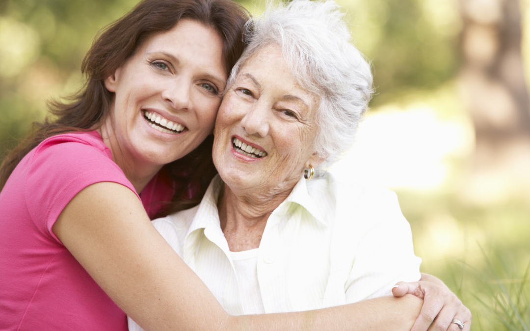 Dark-haired woman in a pink tee shirt embracing an older grey-haired lady in a white shirt. They're both smiling.