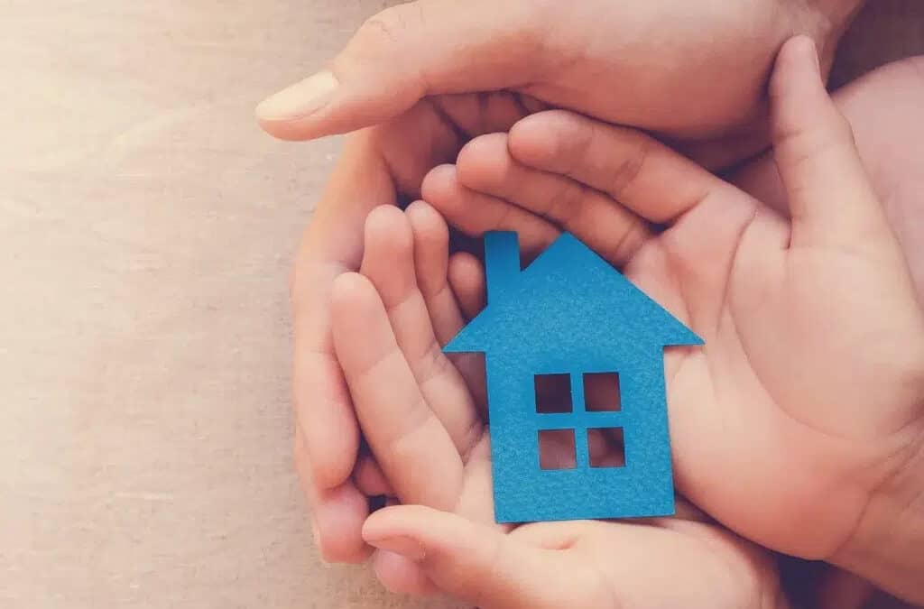 A blue cardboard house being held in a child's hands which are also being held by an adult's hands.