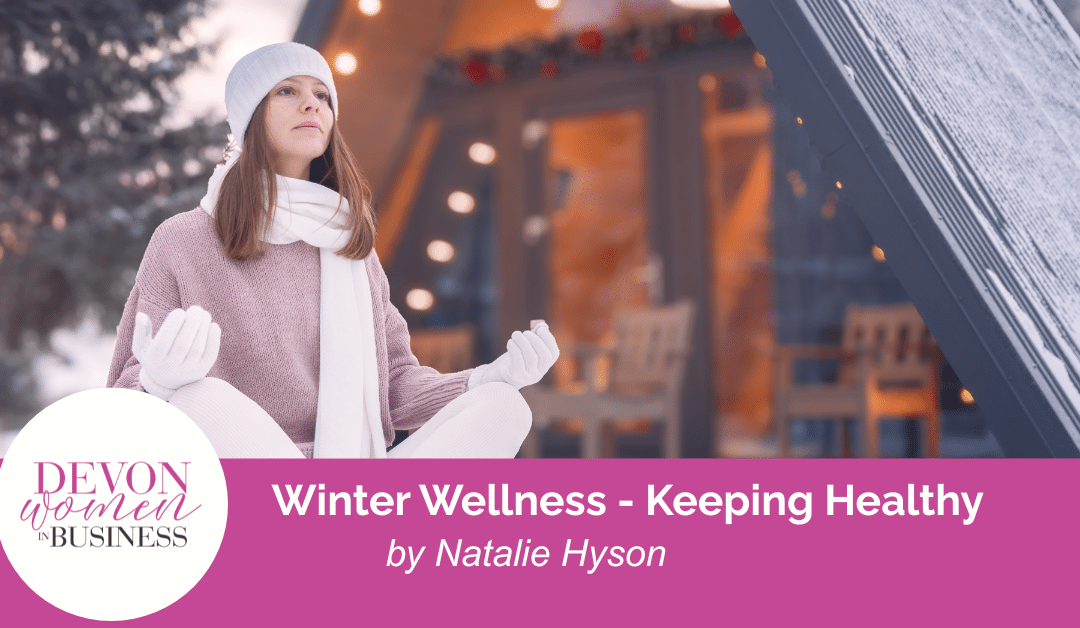 Woman sitting cross-legged in meditation outside a wooden lodge in winter, wearing a hat, scarf, and gloves. Banner text reads “Winter Wellness – Keeping Healthy by Natalie Hyson” with Devon Women in Business logo.