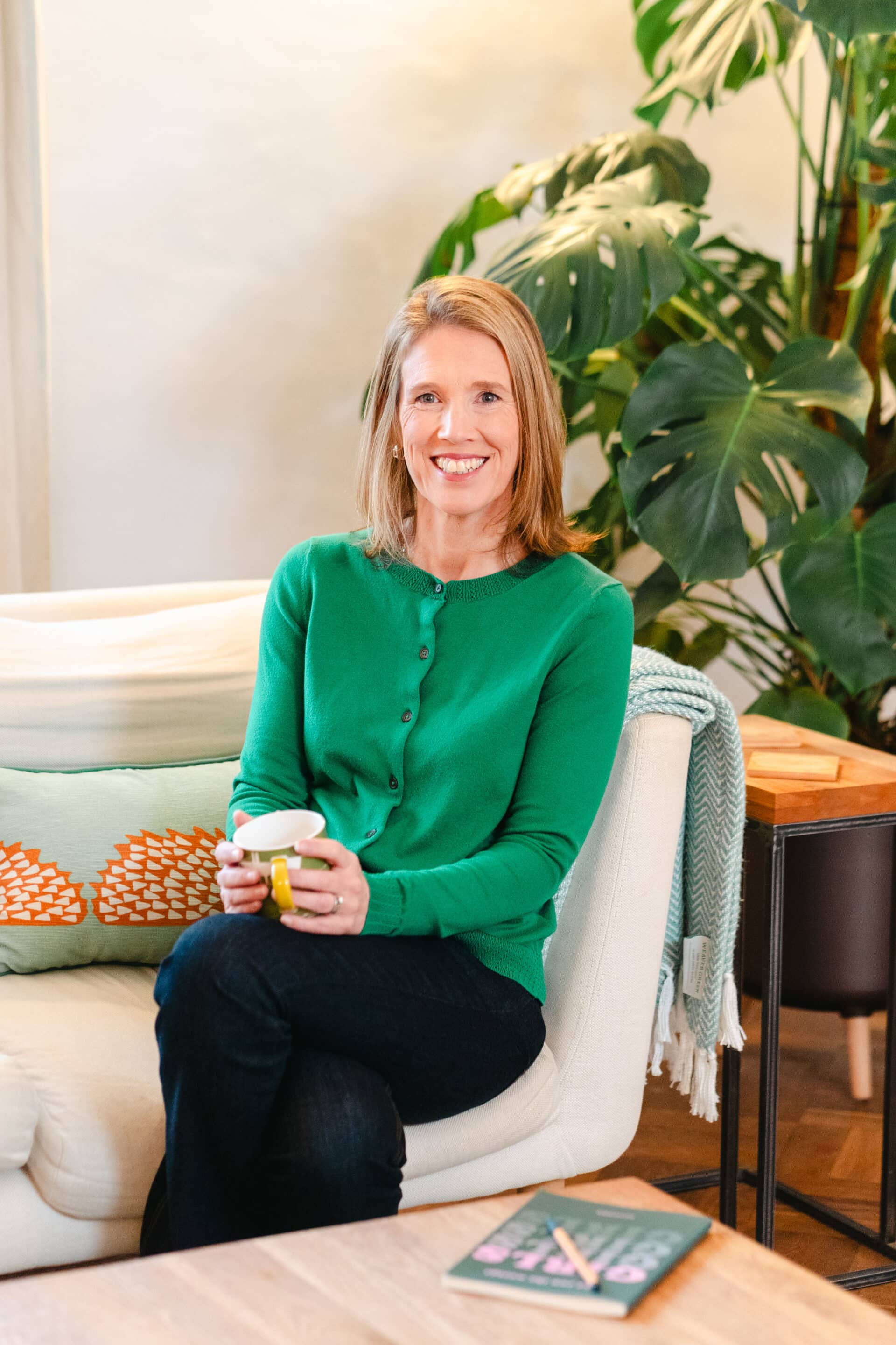 Louise Horrocks Photo of Dr Louise Horrocks sitting on a sofa, She's smiling and holding a mug on her lap. She's wearing a striking green cardigan.
