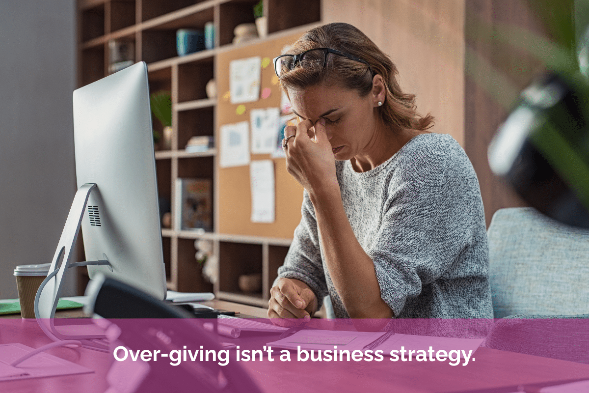 Overwhelmed business owner sitting at a desk with her hand to her forehead beside a computer screen, with the quote “Over-giving isn’t a business strategy.” overlaid in white text on a pink banner.