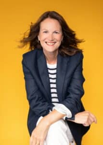 A professional head-and-shoulders portrait of Jackie Williamson smiling warmly at the camera. She is wearing a navy blazer over a white-and-navy striped top with white trousers, and is posed leaning forward with one arm resting across her knee. Her dark brown shoulder-length hair is gently blown back, creating a sense of movement. The portrait is set against a vibrant mustard-yellow background, giving it a bright, confident and approachable feel.