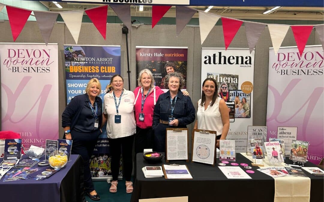 Five women stand together at the Devon Women in Business shared exhibition stand at the Devon Business Show 2026, with banners for Newton Abbot Racecourse, Kirsty Hale Nutrition and Athena Magazine.