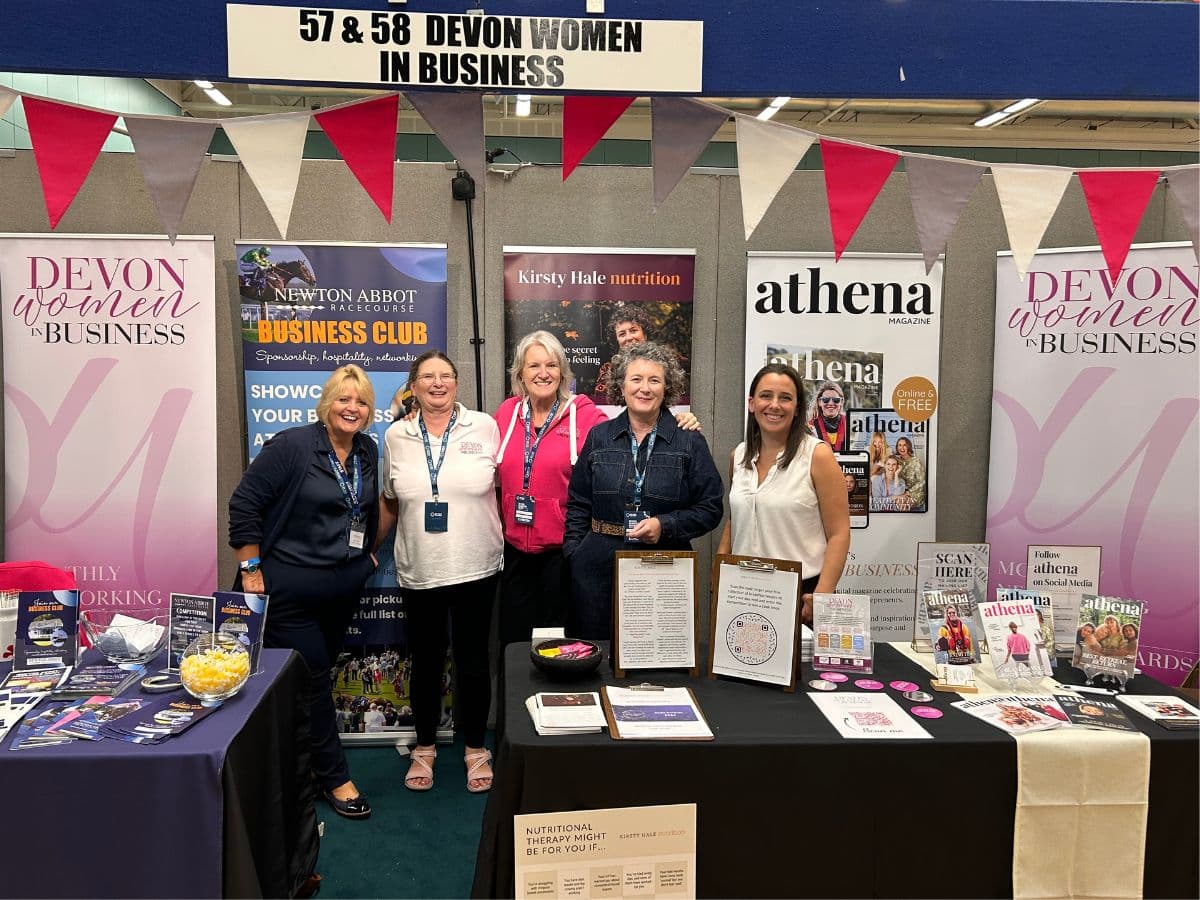 Five women stand together at the Devon Women in Business shared exhibition stand at the Devon Business Show 2026, with banners for Newton Abbot Racecourse, Kirsty Hale Nutrition and Athena Magazine.