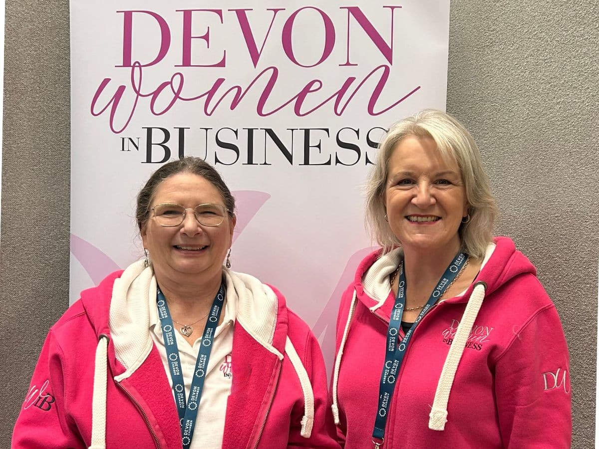Louise Jenner and Maureen Townsend, Co-Leaders of Devon Women in Business, smiling in matching bright pink DWIB hoodies in front of a Devon Women in Business banner.